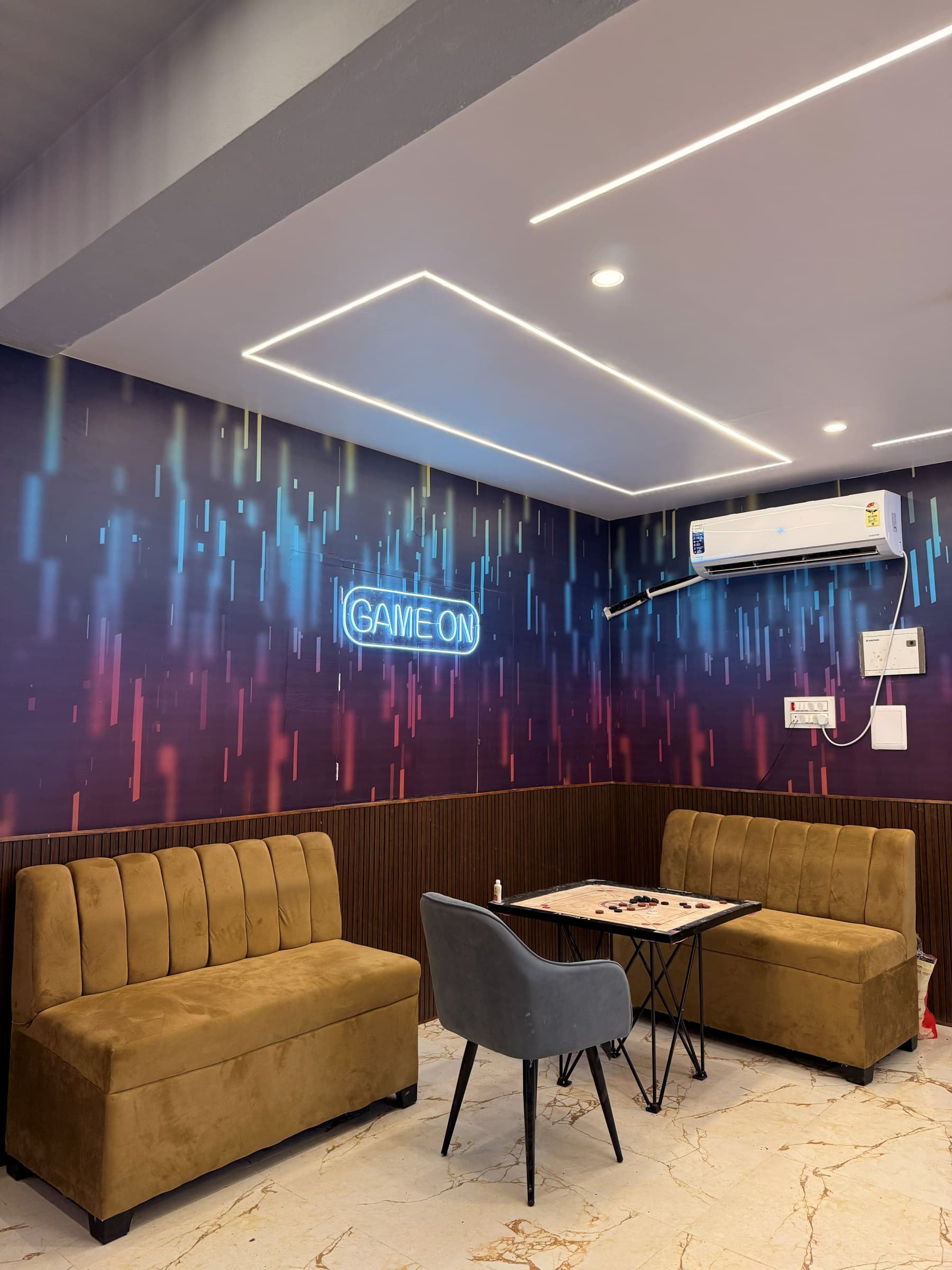 The games lounge with a neon Game On sign, tan sofas and a carrom board